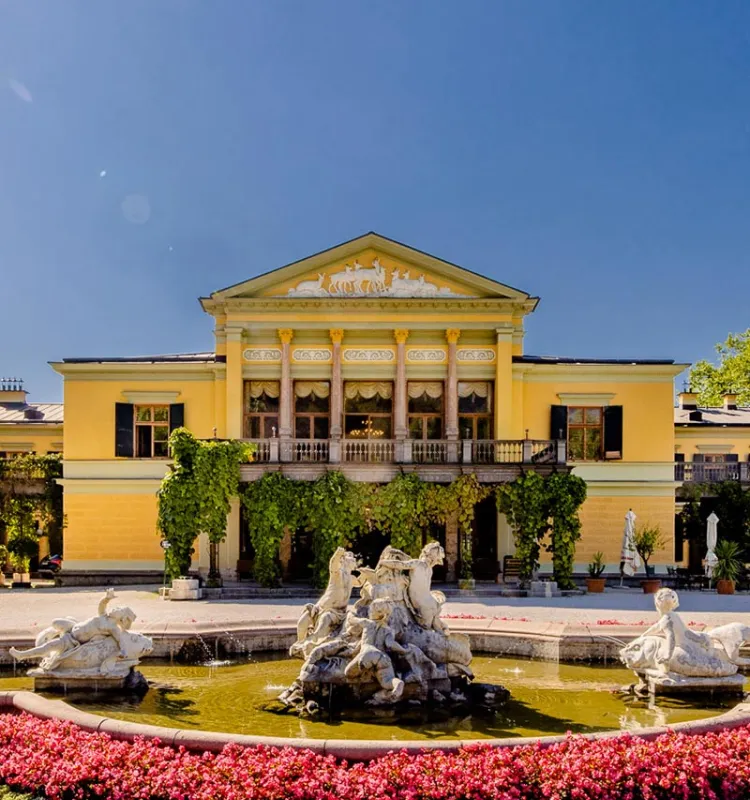 Kaiservilla in Bad Ischl mit Brunnen, Statuen und blühendem Blumenbeet unter blauem Himmel
