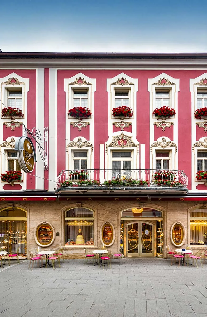 Historische Fassade des Stammhaus der Konditorei Zauner in der Pfarrgasse mit roten Blumen und Außentischen.