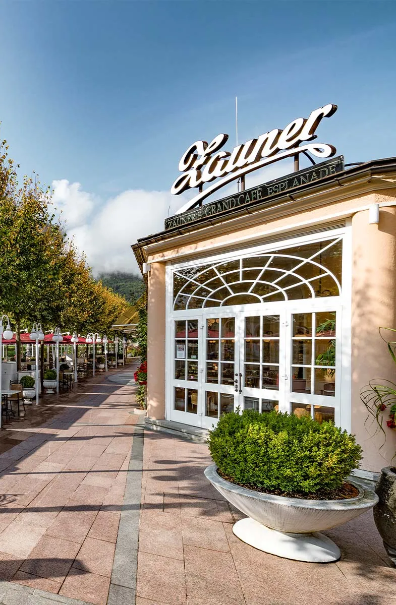 Eingang zum Grand Café Zauner Esplanade mit Terrasse und Bäumen im Hintergrund.