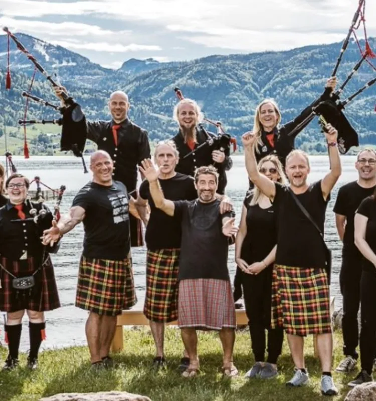 Group of bagpipe players in kilts and black uniform jackets posing cheerfully at the lakeside in the Salzkammergut, with mountains and a calm lake in the background.