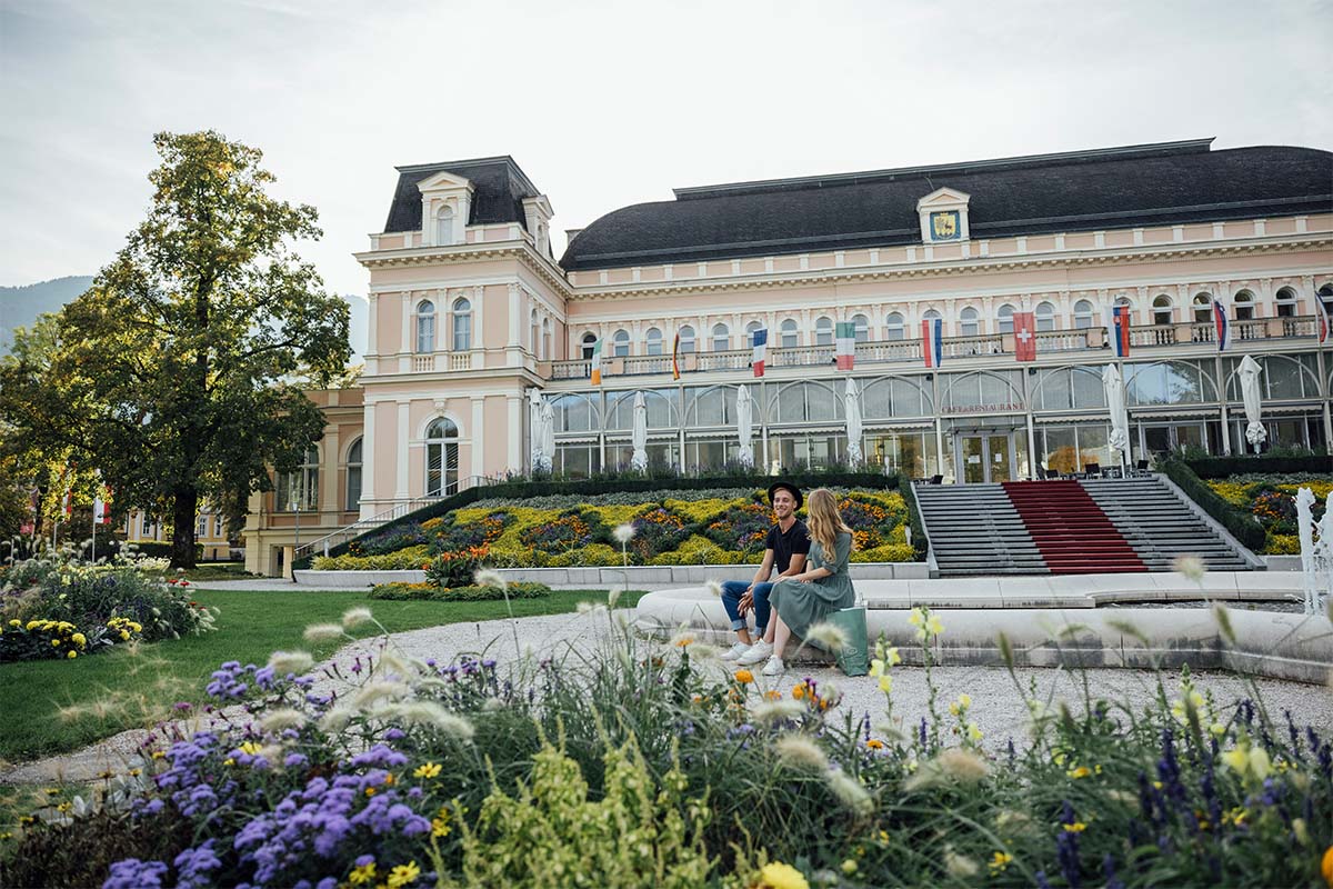 Paar sitzt am Rand eines Brunnens im Park des Theater u. Kongresshauses Bad Ischl nahe dem Hotel Grand Elisabeth